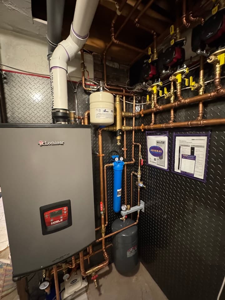 Wall-mounted boiler with copper piping, white vent pipes, an expansion tank and a blue filter housing on a dark backboard.