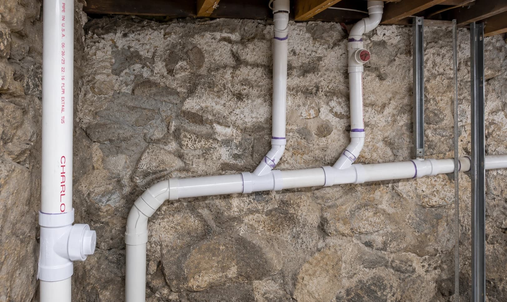White plastic piping with purple-primed joints on a stone basement wall.