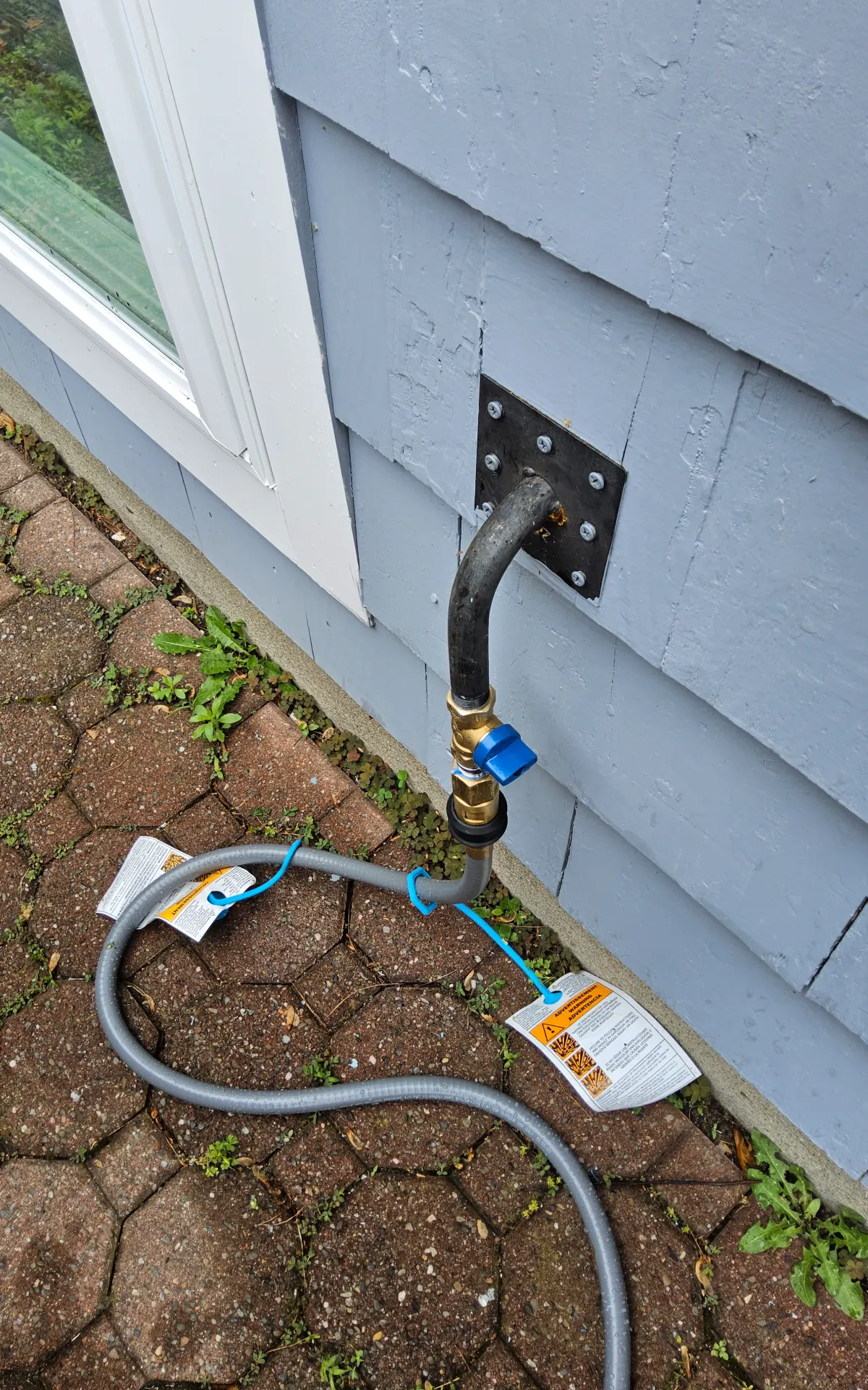 Exterior gas stub through a steel wall plate on shingle siding, with a blue-handled shutoff valve, a quick-disconnect fitting, and a gray flexible gas hose