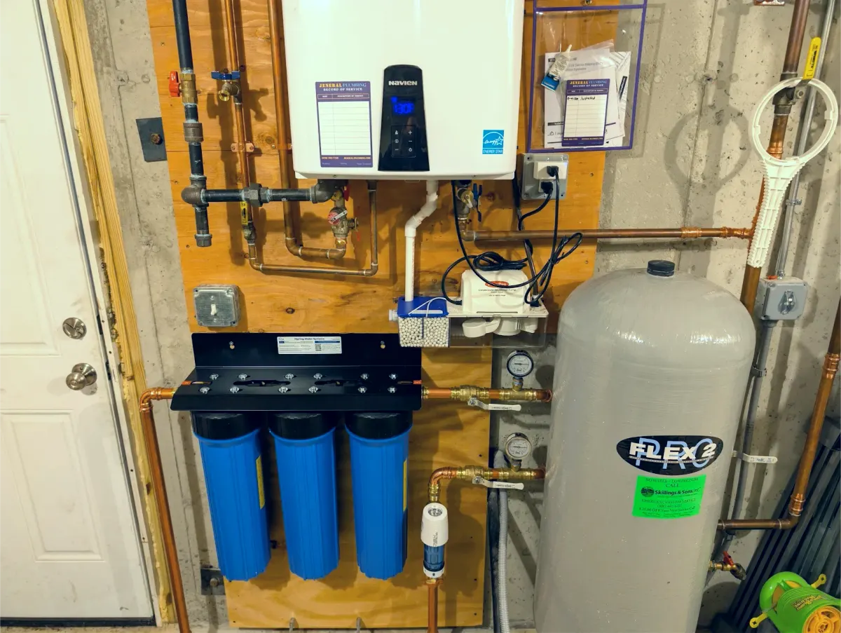 Navien tankless water heater with a Jeneral Plumbing service record label, a three-stage whole-house filter system with pressure gauges, a condensate neutralizer and pump, and a well pressure tank
